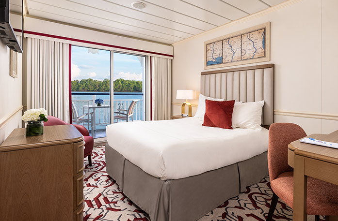 The Single Balcony stateroom on American Heritage, with floor-to-ceiling sliding glass doors that lead to a private balcony with a table and chairs overlooking the Mississippi River. There is a bed on the right side of the room and an end table with a lamp on one side of the bed. There is a wooden dresser and a TV on the wall above it across from the bed, and a red chair in the corner.