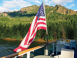 A vibrant American flag flying from the glass railing on the deck of American Jazz, an American Riverboat small cruise ship while sailing along the Columbia River. Alongside the river, there is a cliff with thick forests spanning up the side.