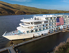 American Jazz, an American Riverboat small cruise ship docked on a small walkway along the Columbia River. There is a large American flag on the side of the cruise ship, and there are small, grassy hills on the shore on the other side of the river.