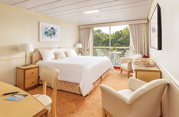 The Premium Balcony stateroom on American Song with floor-to-ceiling glass doors that lead to a balcony with a table and chairs overlooking the Columbia River. There is a bed on the left of the room with pillows and a bed frame with an orange and white pattern. There are two end tables with lamps, two accent chairs with a wood dresser between them, an orange patterned rug, and a TV on the wall.