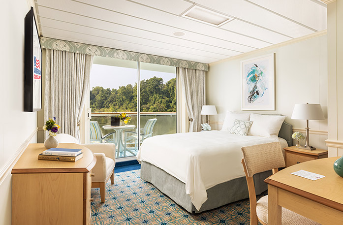 The Single Balcony stateroom on American Song with floor-to-ceiling glass doors that lead to a balcony with a table and chairs overlooking the Columbia River. There is a bed to the right with pillows and a grey bed frame. There are two end tables with lamps, an accent chair in the left corner, a wood dresser under a TV on the wall, and a desk and chair to the right of the bed.