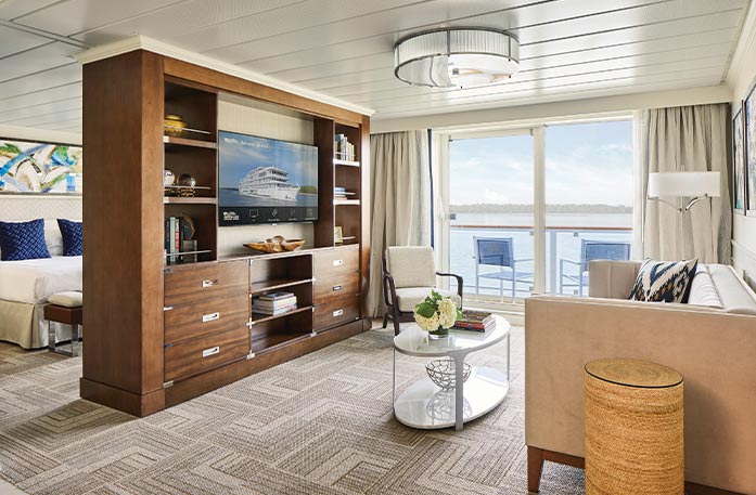 The Owner's Suite on American Song, with floor-to-ceiling glass doors that lead to a balcony with a table and chairs overlooking the Columbia River. There is a tan sofa with a black pillow beside a small table, and a large, wooden TV stand with six drawers and shelves filled with books and decor and a TV in the middle. Behind the tv stand is a bed with dark blue pillows with a painting above