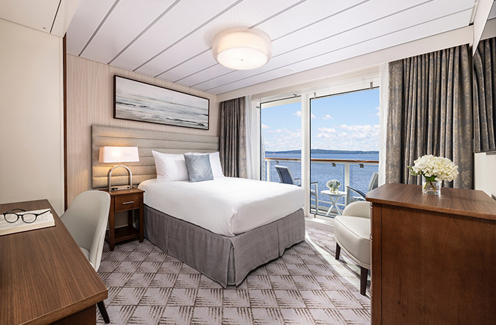 The Single Balcony stateroom on American Legend, with floor-to-ceiling sliding glass doors that lead to a balcony with a table and chairs overlooking the Gulf Coast. here is a bed on the left side of the room with a grey accent pillow, a bedside table with a lamp, and a painting of waves above it. There is a wooden dresser on the right side, and a desk and chair in the foreground. 