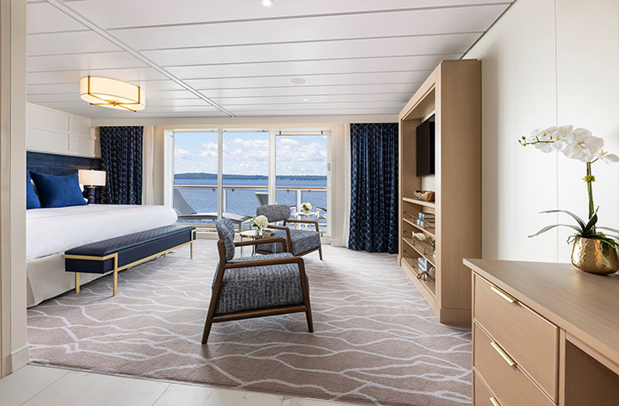 The Sky Suite on American Legend with floor-to-ceiling sliding glass doors that lead to a balcony with a table and chairs overlooking the Gulf Coast. On the left side of the room, there is a bed with blue pillows and a blue bench with gold legs at the foot of the bed. There are two chairs with blue cushions in front of a wooden TV stand with shelves and a TV on it. 