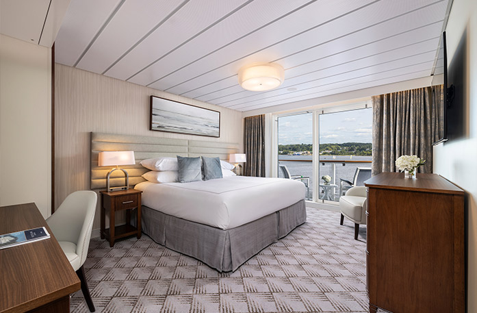 The Vista Balcony stateroom on American Legend with floor-to-ceiling sliding glass doors that lead to a balcony with a table and chairs overlooking the Gulf Coast. There is a bed on the left side of the room with two grey pillows, bedside tables with a lamp on each side, and a painting of waves above it. There is a wooden dresser on the right side of the room with a TV on the wall above it. 