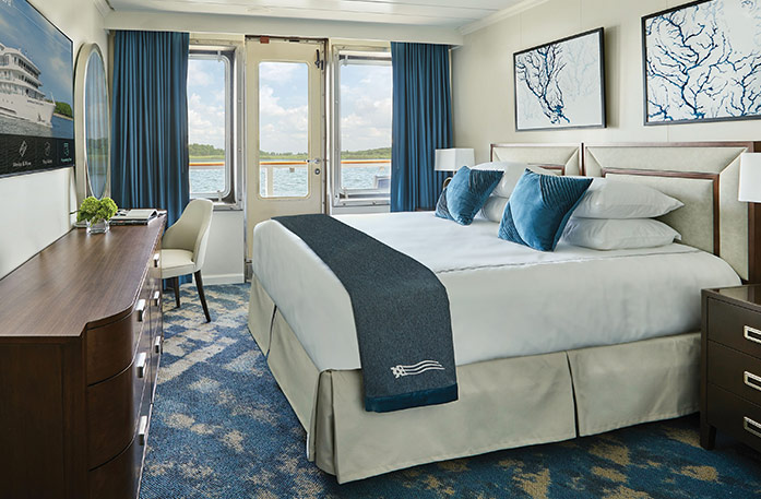 The Standard Balcony stateroom on American Symphony, with a push door with blue curtains that lead to a balcony overlooking the Mississippi River, a bed with white bedding and two blue pillows and a blue bed runner, and two white paintings with blue lines above the bed. There is an end table with a lamp on each side of the bed, and a dark wood dresser and a TV on the wall in front of the bed.