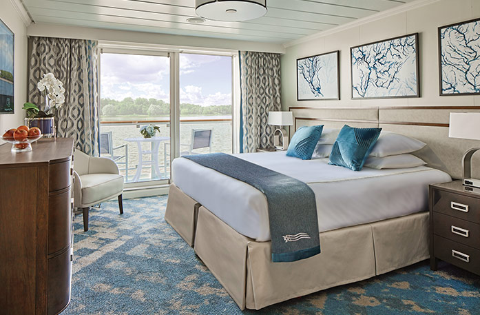 The Deluxe Balcony stateroom on American Symphony, with large floor-to-ceiling glass doors with tan patterned curtains on each end that lead to a balcony with chairs overlooking the Mississippi River, a bed with white bedding and two blue pillows and a blue bed runner, and three white paintings with blue lines above the bed. There is an end table with a lamp on each side of the bed. 