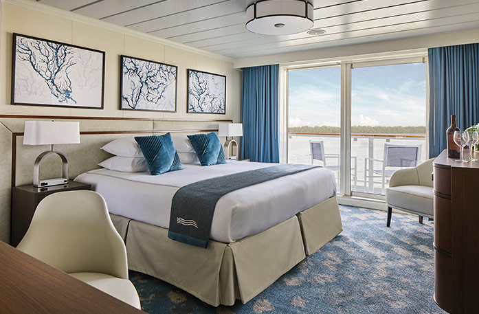 The Premium Balcony stateroom on American Symphony, with large floor-to-ceiling glass doors with blue curtains on each end that lead to a balcony with chairs overlooking the Mississippi River, a bed with white bedding and two blue pillows and a blue bed runner, and three white paintings with blue lines above the bed. There is an end table with a lamp on each side of the bed. 