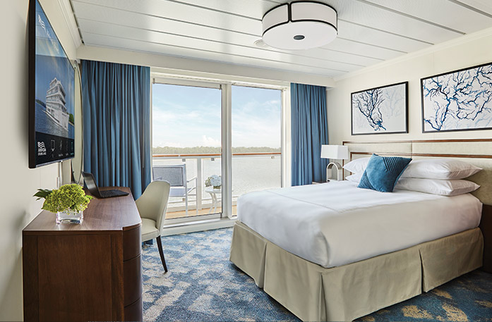 The Single Balcony stateroom on American Symphony, large floor-to-ceiling glass doors with blue curtains on each end that lead to a balcony with a chair and table overlooking the Mississippi River, a bed with white bedding and one blue pillow, and two white paintings with blue lines above the bed. There is a dark wood dresser and a TV on the wall in front of the bed.