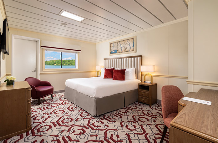 The Standard Balcony stateroom on American Pride, with floor-to-ceiling sliding glass doors that lead to a private balcony with a table and chairs overlooking the Columbia River. There is a bed on the left side of the room and end tables with a lamp on each side of the bed, across from a wooden dresser and a TV on the wall above it across from the bed, and a red chair in the corner.