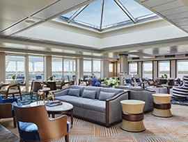 The Sky Lounge, a common area aboard American Serenade, with windows looking out to the Mississippi River on both walls pictured, several round tables with blue and brown chairs beside them, and two grey sofas in the center. 