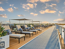 Several white lounge chairs with blue patterned pillows are placed next to a large plant in a grey, square pot along the sky walk on the American Serenade sun deck.