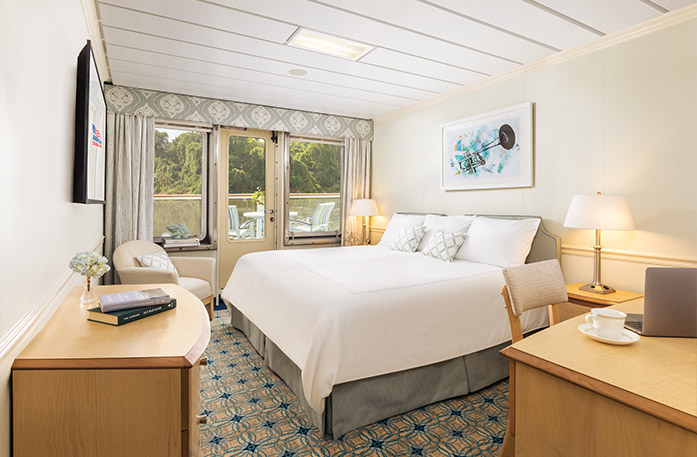 The Standard Balcony 2 stateroom on American Song with a push door that leads to a balcony with a table and chairs overlooking the Columbia River. There is a bed to the right with pillows and a grey bed frame. There are two end tables with lamps, an accent chair in the left corner, a wood dresser under a TV on the wall, and a desk and chair to the right of the bed.