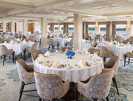 The Restaurant aboard American Splendor Paddlewheeler, where there are several separate tables draped with white table cloths with folded napkins, silverware, glassware, and a vase with flowers on each. Each table has a few plush chairs surrounding it. The back wall features several windows that provide natural lighting and a view of the Mississippi River.