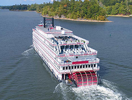 American Heritage, an American Cruise Lines Paddlewheeler decorated with patriotic red white and blue bunting flags on the back railings, sailing along the Mississippi River. The back of the small cruise ship has a large, red paddlewheel, and the shore on the right side of the river is covered in trees.