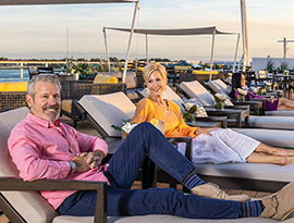 Three cruise guests lay on lounge chairs on the Sun Deck on American Symphony, while a couple is holding hands while walking on the Sun Walk, a blue walking trail along glass railings.