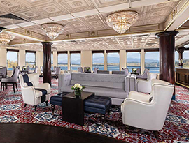 The River Lounge, a common area aboard American West, an American Cruise Lines Paddlewheeler. The lounge has a grey sofa in the center with two white leather chairs beside it, and an ottoman with a small table in the center. The walls contain many large windows that provide the perfect view of the Columbia River.