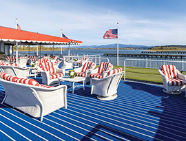 The sun deck on American West, an American Cruise Lines Paddlewheeler, where there are several red and white striped chairs paired with small, white round tables. The floor of the sun deck is blue with small white stripes, and there is a putting green along the railing, where American flags fly, with views of the Columbia River and mountains lining the horizon in the distance.