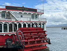 A close-up view of the back of American West, an American Cruise Lines Paddlewheeler small cruise ship sailing on the Columbia River. There is a large, red paddlewheel attached to the center of the backside of the ship, and "American West" written in gold letters on a black backdrop on the railing of the top deck. In the distance, there are silhouettes of mountains and thick clouds in the sky.