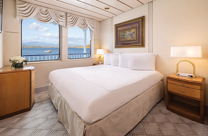 The Standard Waterview stateroom on American West, with two large windows overlooking the Columbia River. There is a bed on the right side of the room and end tables with a lamp on each side of the bed, and a wooden dresser across from the bed in the corner.