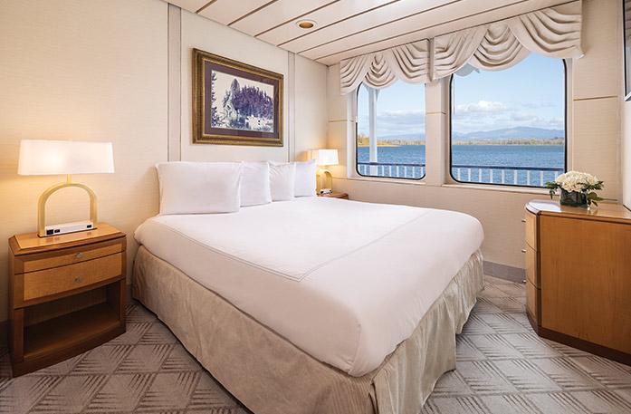 The Deluxe Waterview stateroom on American West, with two large windows overlooking the Columbia River. There is a bed on the left side of the room and end tables with a lamp on each side of the bed, and a wooden dresser across from the bed in the corner.