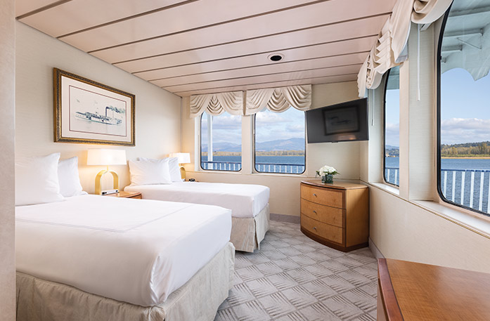 The Premium Waterview stateroom on American West, with four large windows overlooking the Columbia River. There are two beds on the left side of the room with end tables with a lamp, and in the corner of the room, there is a wooden dresser and a TV on the wall above it.