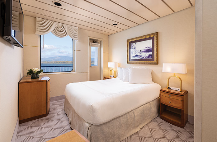 The Single Balcony stateroom on American West, with a push door that leads to a private balcony, beside a large picture window overlooking the Columbia River. There is a bed on the right side of the room, and an end tables with a lamp on each side of the bed. There is a wooden dresser in the corner and a TV on the wall above it across from the bed.